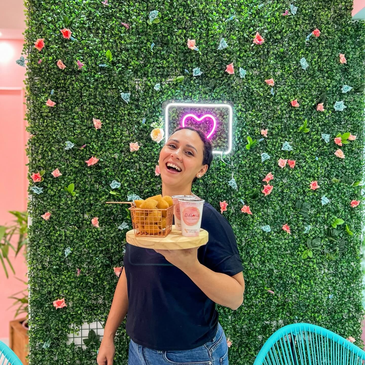 A Débi segurando uma bandeja de salgados em frente à parede de folhagem com neon de coração